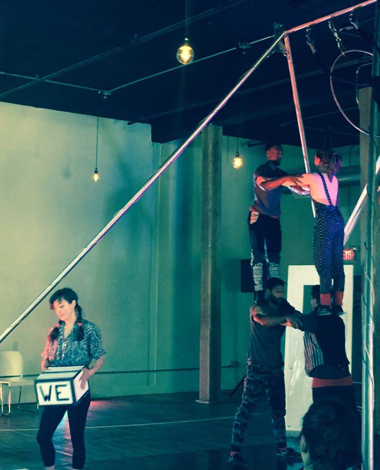 two people standing on an additional two people's shoulders with arms intertwined. Separate person holds a box that says "we".